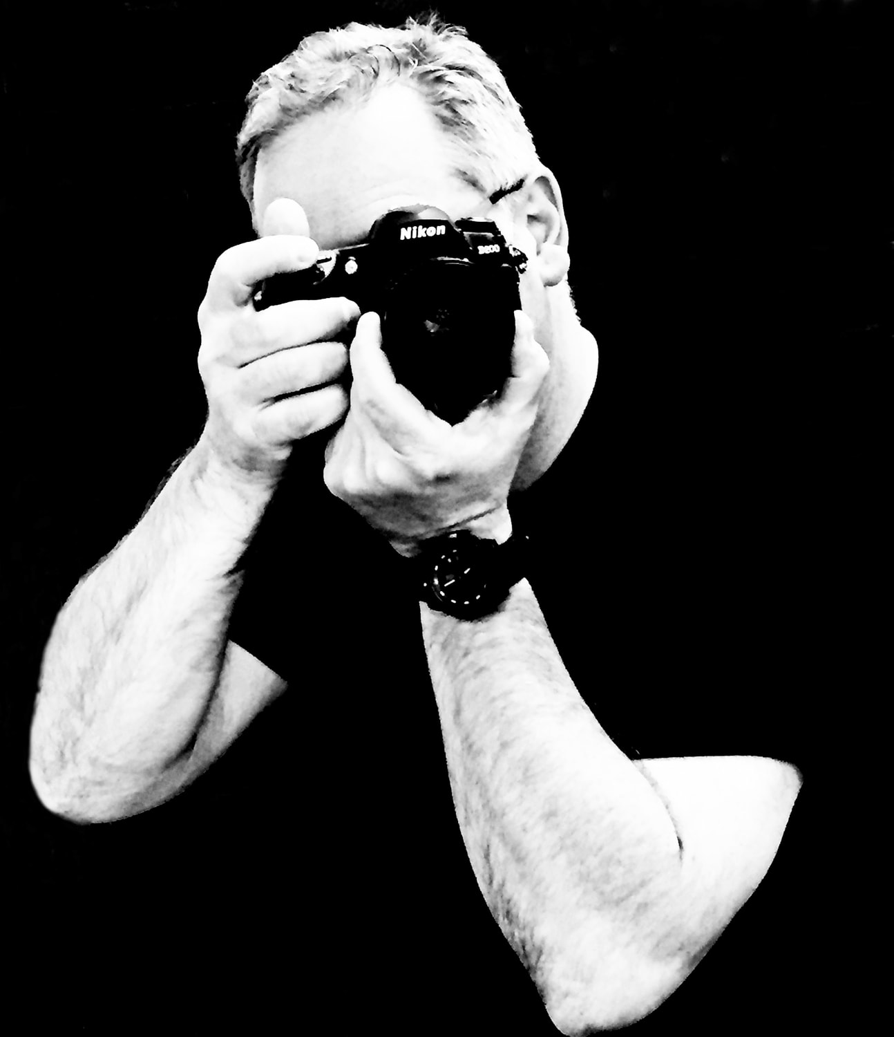 Black-and-white portrait of Rob Conens holding a Nikon camera to his face against a black ground.